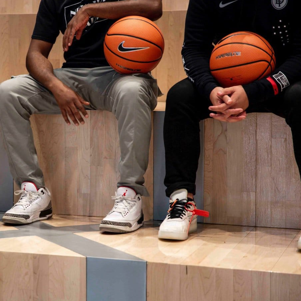 Modelos sentados mostrando dos modelos de tenis de la marca nike, además sujetan un balón naranja de basquetbol con el logo de la marca