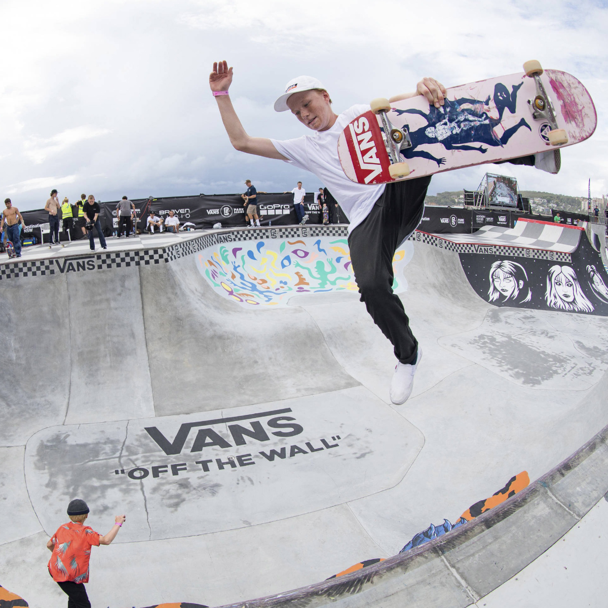 Persona en parque de skate realizando un truco en el aire con su tabla de patinaje, la cual es blanca con el logotipo de Vans en color rojo, de fondo personas observan cómo se realiza el truco